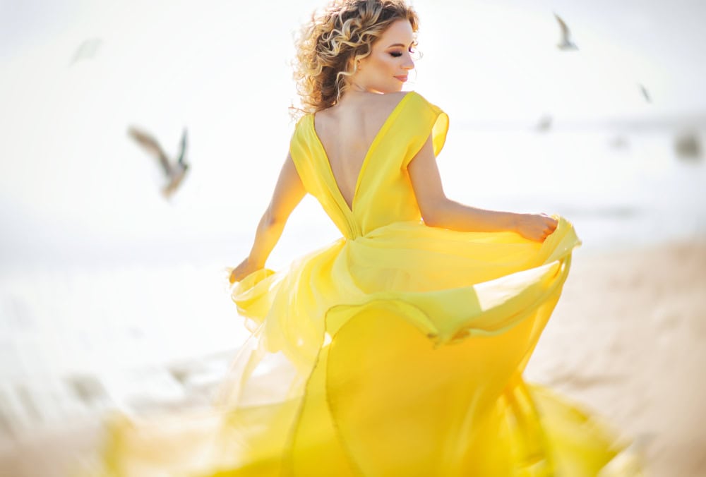 Woman in bright yellow dress walking on the beach with birds flying around, captured in soft sunlight