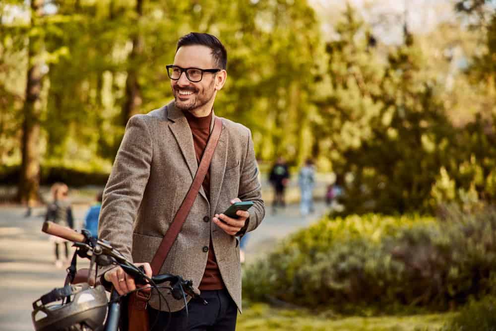 Smiling man in a blazer holding a phone and standing with his bike in a sunlit park