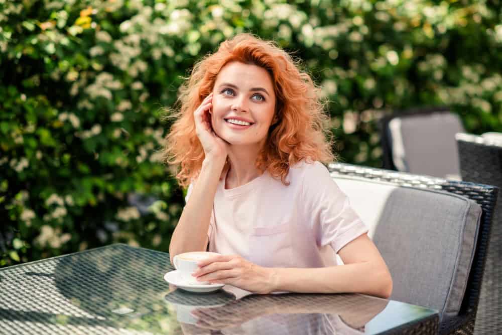 Smiling female model with curly red hair drinking coffee at an outdoor cafe in the sunshine